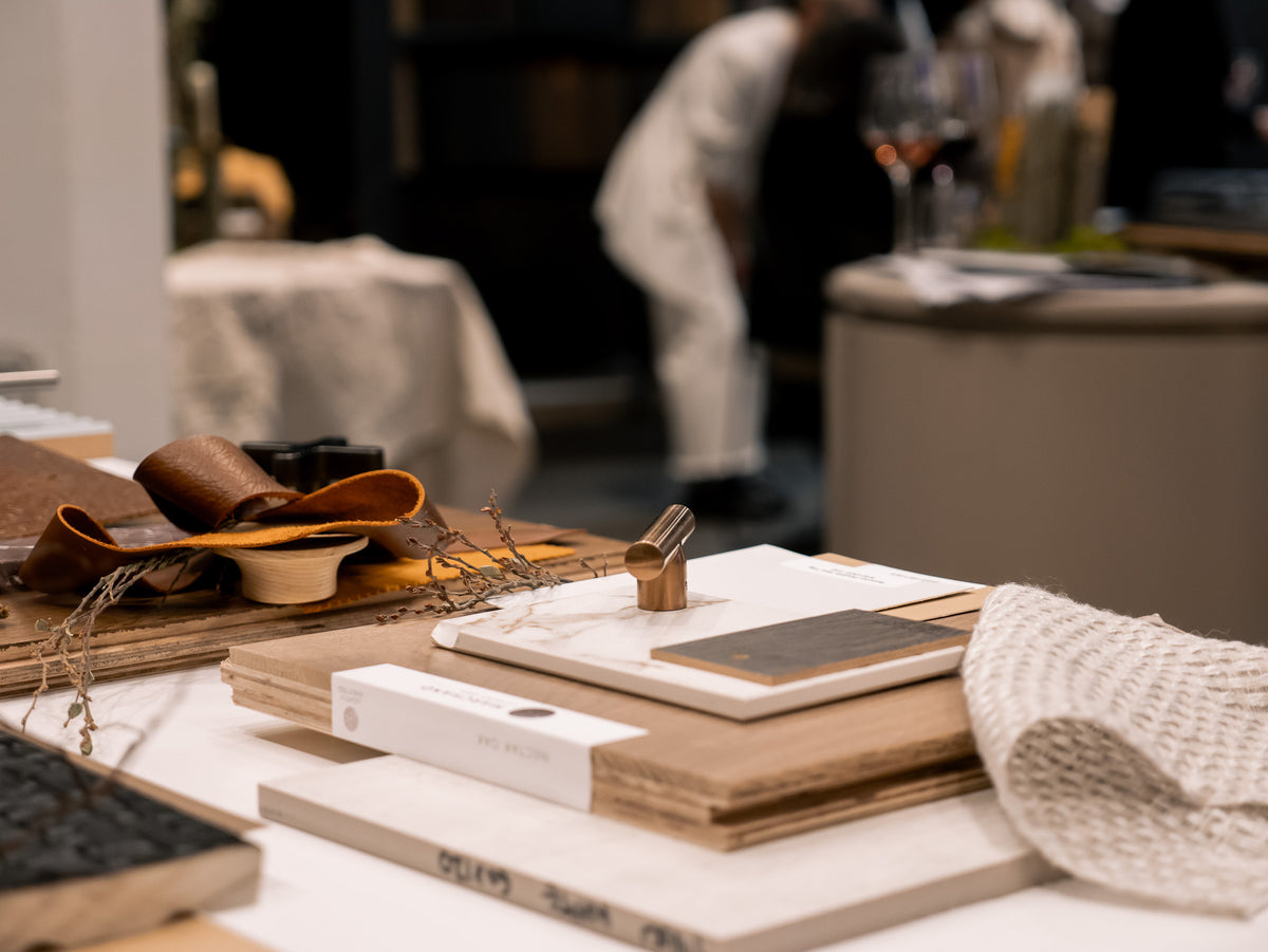 Material flatlay in the Marchand studio: pale European oak sample boards stacked with leather, linen and stone swatches, the studio busy behind