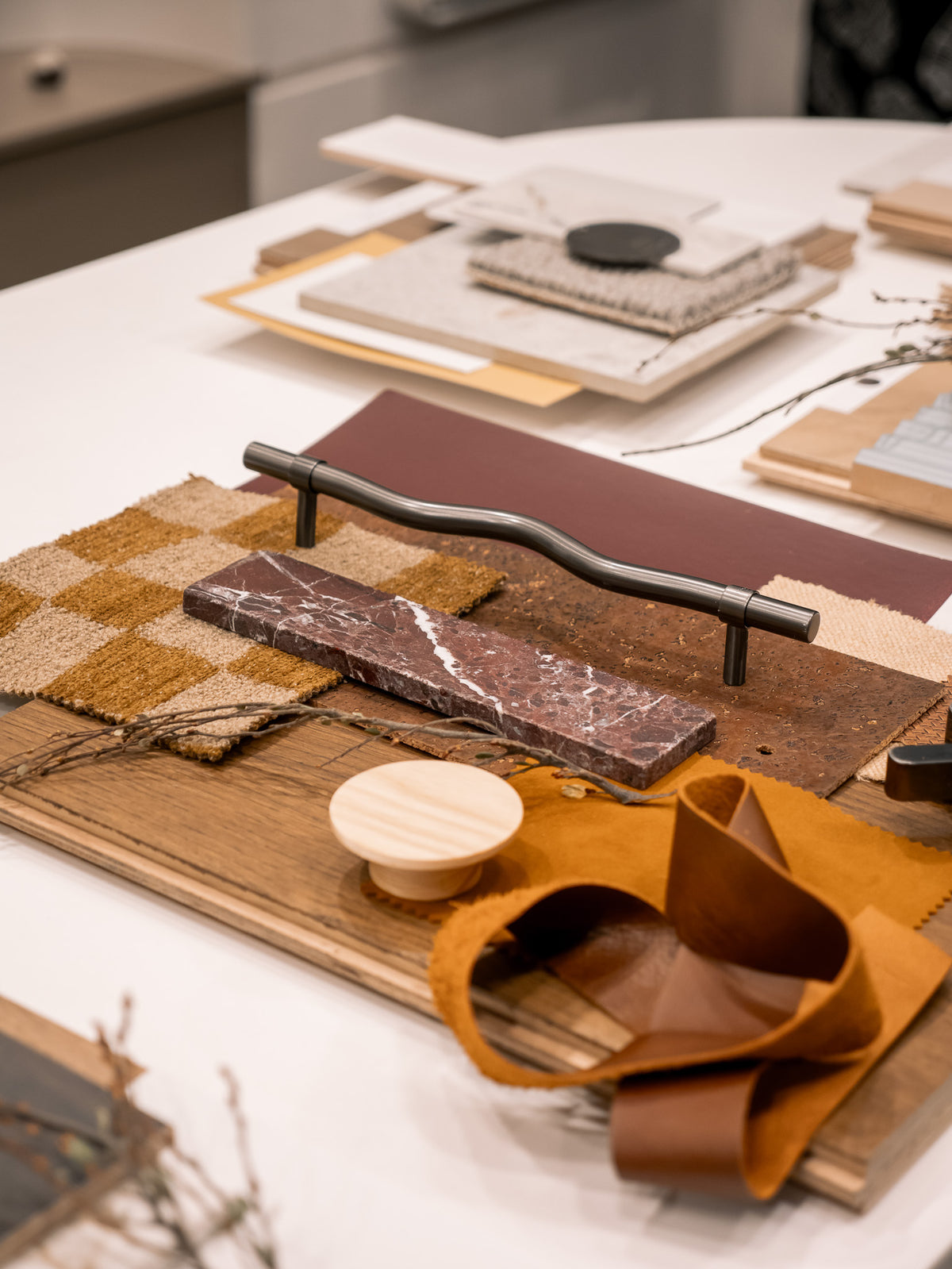Warm Minimal material flatlay in the Marchand studio: warm European oak sample board with tan leather, a burgundy swatch, checked textile, a black handle and a timber stool in the background